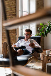 Fokussierter männlicher Unternehmer, der in einem bequemen Sessel sitzt und sich Notizen macht, während er seinen Arbeitstag im Home Office plant - ADSF23630