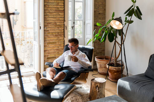 Seriöser Geschäftsmann, der zu Hause im Sessel sitzt und Pläne in einen Organizer schreibt, während er in der Pause einen Kaffee genießt - ADSF23631