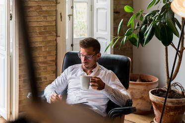 Seriöser Geschäftsmann, der zu Hause im Sessel sitzt und Pläne in einen Organizer schreibt, während er in der Pause einen Kaffee genießt - ADSF23632