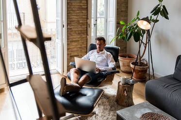Gut aussehender männlicher Unternehmer, der zu Hause im Sessel sitzt und an einem Projekt auf dem Laptop arbeitet, während er einen Kaffee genießt - ADSF23637