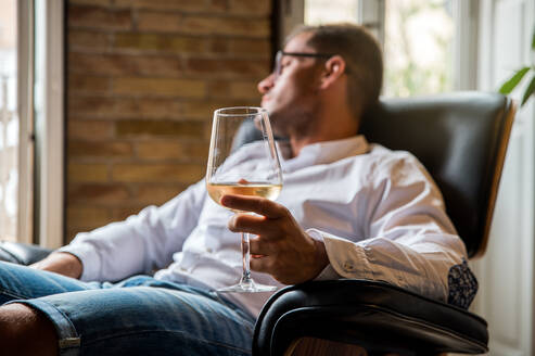 Pensive male resting in comfortable leather armchair with glass of white wine and looking away in thoughts - ADSF23640