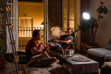 Relaxed men sitting in cozy living room and smoking hookah together while enjoying evening at weekend - ADSF23658