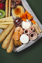 Von oben Brunch-Box mit verschiedenen Arten von Käse und Knäckebrot in der Nähe von reifen Tasse Kiwi süße Erdbeeren und geschälte Mandarine in der Nähe von Marmelade in Glas auf bunten grünen Hintergrund angeordnet geschnitten Fleisch - ADSF23660