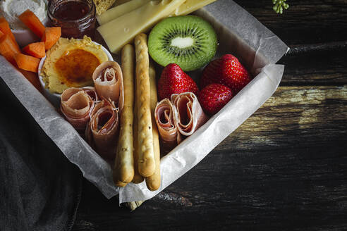 Von oben Brunch-Box mit verschiedenen Aufschnittarten verschiedene Arten von Käse und Knäckebrot in der Nähe von reifen Becher Kiwi süße Erdbeeren und geschälte Mandarine in der Nähe von Marmelade im Glas auf Holztisch in der Nähe Serviette angeordnet - ADSF23663