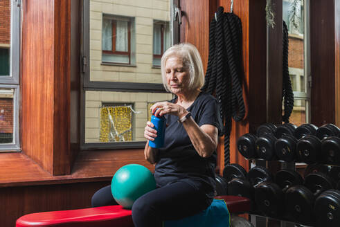 Ältere Sportlerin, die eine Flasche öffnet, während sie auf einer Bank mit einem kleinen Gymnastikball gegen Hanteln in einer Sporthalle sitzt - ADSF23666