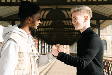 Seitenansicht von stilvollen jungen diverse männliche Freunde in trendigen Outfits stehen auf gepflasterten Straße in der Stadt und schütteln die Hände - ADSF23686