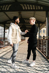 Side view of stylish young diverse male friends in trendy outfits standing on paved street in city and shaking hands - ADSF23687