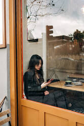 Langhaarige brünette asiatische Frau, die in einem Café einen Kaffee trinkt, während sie ein Handy betrachtet - ADSF23690