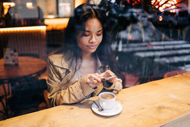 Long-haired brunette Asian woman having a coffee on a coffee shop while is looking a cellphone - ADSF23709
