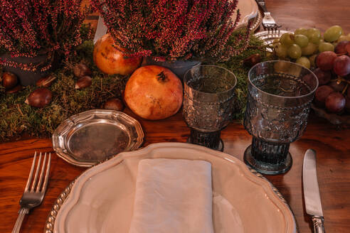 Hohe Winkel der Kristallgläser in der Nähe von Teller und Besteck auf dem Tisch mit Trauben Calluna vulgaris Blumen und Granatapfel dekoriert platziert - ADSF23731