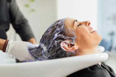 Crop male hairdresser in apron washing hair of female client in sink after cutting and dyeing in modern beauty salon - ADSF23759