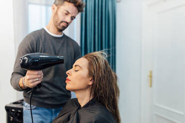 Crop junge ethnische männliche Friseur Trocknen Haare der weiblichen Kunden mit geschlossenen Augen in der modernen Schönheit Studio - ADSF23760