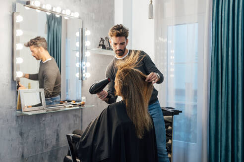 Ethnischer männlicher Friseur trocknet die Haare einer Kundin mit geschlossenen Augen in einem modernen Schönheitsstudio - ADSF23761