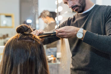 Ein männlicher Stylist in Freizeitkleidung benutzt ein Glätteisen, während er einer Kundin im Schönheitssalon Locken macht - ADSF23762