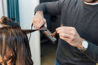 Crop unrecognizable male stylist in casual clothes using hair straightener while doing curls for female client in beauty salon - ADSF23763