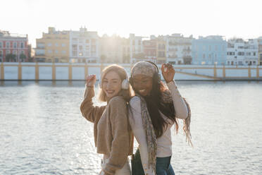 Seitenansicht von multirassischen Freundinnen mit Kopfhörern, die an einem sonnigen Tag Musik genießen und zusammen am Ufer tanzen - ADSF23767