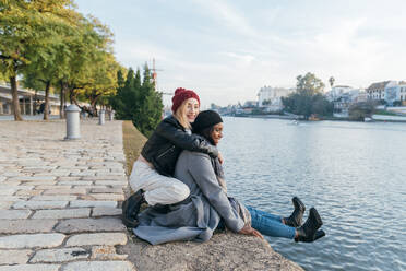 Seitenansicht von fröhlichen multiethnischen Freundinnen, die sich auf der Promenade umarmen, während sie ein gemeinsames Wochenende im Herbst genießen und wegschauen - ADSF23771