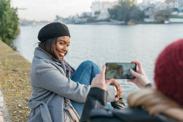 Unrecognizable woman taking picture of black female friend on smartphone while relaxing on embankment in city - ADSF23773