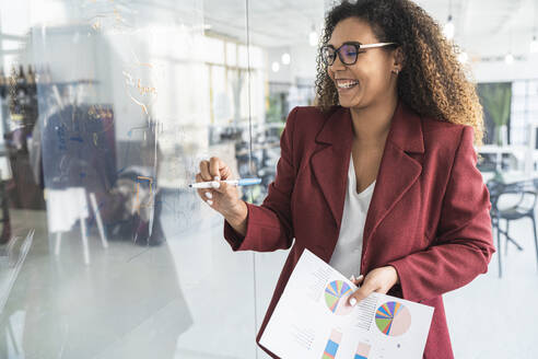 Smiling businesswoman holding financial report while writing on glass in office - SNF01279