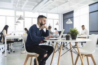 Männliche Fachkraft arbeitet an einem Laptop mit Kollegen im Hintergrund in einem Coworking-Büro - SNF01305