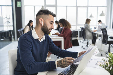 Ein männlicher Unternehmer liest ein Dokument, während er vor einem Laptop sitzt und mit männlichen und weiblichen Kollegen in einem Coworking-Büro arbeitet - SNF01309