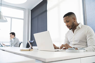 Geschäftsmann arbeitet am Laptop mit einem männlichen Kollegen im Hintergrund in einem Coworking-Büro - SNF01317