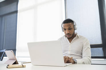 Male entrepreneur wearing headphones working on laptop at office - SNF01318