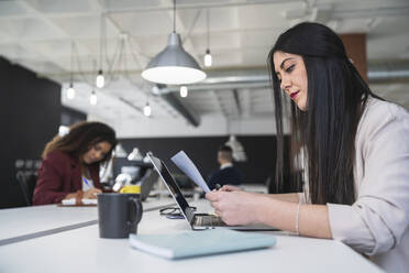 Geschäftsfrau, die ein Dokument liest, während im Hintergrund eine Kollegin in einem Coworking-Büro arbeitet - SNF01328