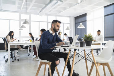 Männlicher Unternehmer arbeitet am Laptop mit Kollegen im Hintergrund in einem Coworking-Büro - SNF01336