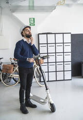 Male entrepreneur removing helmet while standing with push scooter arriving in office - SNF01340