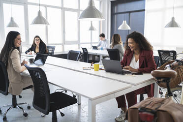 Lächelnde Unternehmerinnen, die am Schreibtisch in einem Coworking-Büro diskutieren - SNF01348