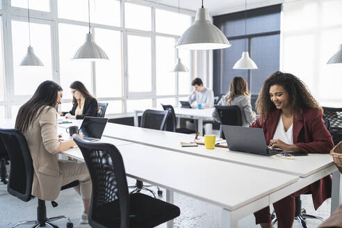 Female entrepreneurs smiling while using laptop with colleagues in background at office - SNF01349