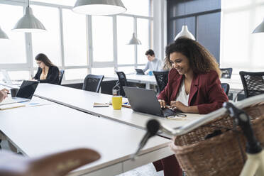 Lächelnde Geschäftsfrau arbeitet am Laptop mit männlichen und weiblichen Kollegen im Hintergrund im Büro - SNF01359