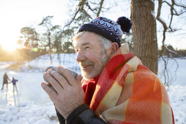 In eine Decke eingewickelter älterer Mann, der im Winter mit einer Tasse in der Hand wegschaut - FVDF00130