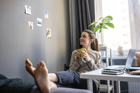 Lächelnde Frau mit Kaffeetasse träumt, während sie ein Foto an der Wand betrachtet - VPIF03915