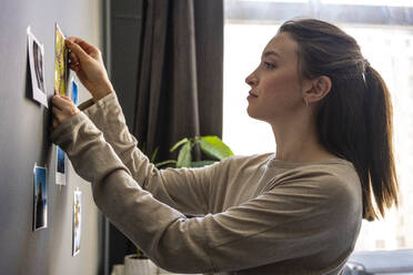 Young woman sticking photograph on wall at home - VPIF03919