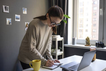 Young woman writing in book while standing by laptop at home - VPIF03923