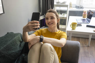 Lächelnde Frau nimmt Selfie durch Smartphone auf Sofa im Wohnzimmer - VPIF03931