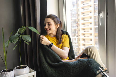 Woman touching leaf while sitting by window - VPIF03935