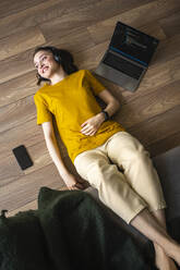 Smiling woman with headphones lying by laptop on floor - VPIF03945