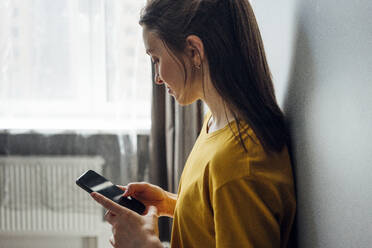 Woman using mobile phone while leaning on wall at home - VPIF03954