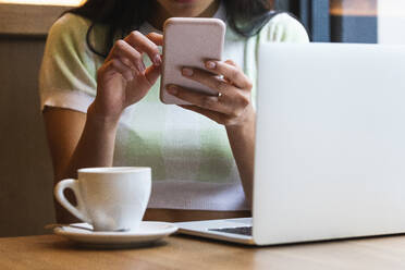 Frau mit Laptop und Kaffeetasse, die ihr Smartphone in einem Café benutzt - PNAF01560