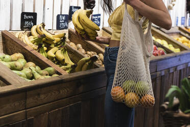 Junge Frau mit Netzbeutel beim Kauf von Bananen im Lebensmittelgeschäft - PNAF01567