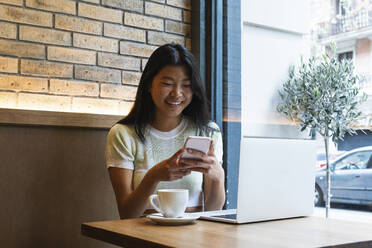 Lächelnde Frau, die ihr Smartphone am Laptop benutzt, während sie in einem Café sitzt - PNAF01576