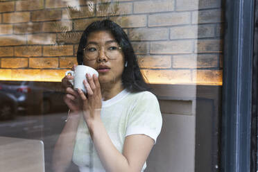 Junge Frau mit Kaffeetasse schaut durch Glas in einem Café - PNAF01580