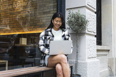 Woman using laptop while sitting on bench outdoors - PNAF01582
