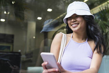 Woman in bucket hat using smart phone by glass wall - PNAF01597