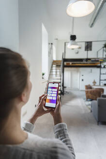 Junge Frau, die zu Hause über ihr Mobiltelefon auf elektrische Geräte zugreift - MCVF00777