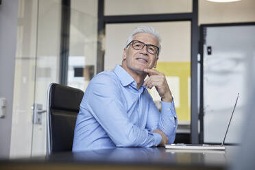 Thoughtful businessman sitting with hand on chin by laptop at office - RBF08076