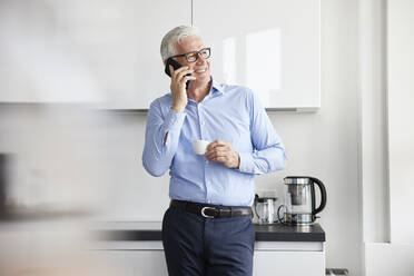 Lächelnder Geschäftsmann mit Tasse, der in einer Cafeteria mit seinem Mobiltelefon spricht - RBF08095